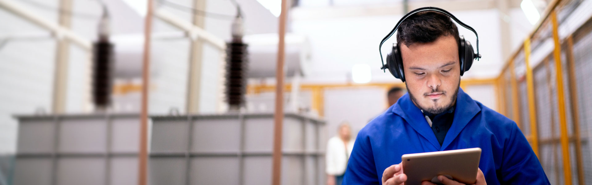 Young man with Down syndrome wearing a royal blue shirt and headphones stands in a warehouse while working on a tablet.